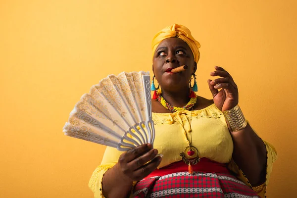 Mulher americana africana de meia idade em turbante amarelo e roupas brilhantes segurando fã e fumar charuto no amarelo — Fotografia de Stock