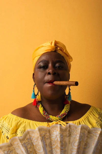 Portrait of serious middle aged african american woman smoking cigar on yellow — Stock Photo