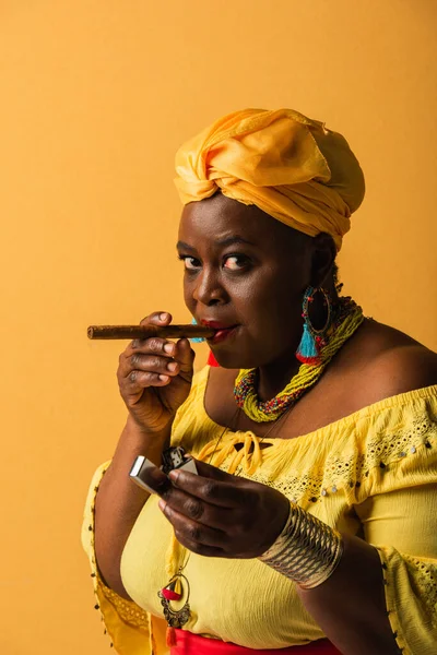 Середнього віку афроамериканська жінка в жовтому тюрбані і блузка дивиться на камеру з запальничкою і сигарою в руках на жовтому — стокове фото