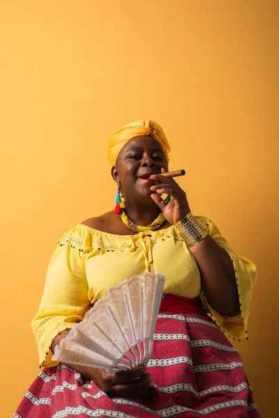 Середнього віку афроамериканська жінка в яскравому одязі, сидячи з вентилятором і палити сигару на жовтому — стокове фото