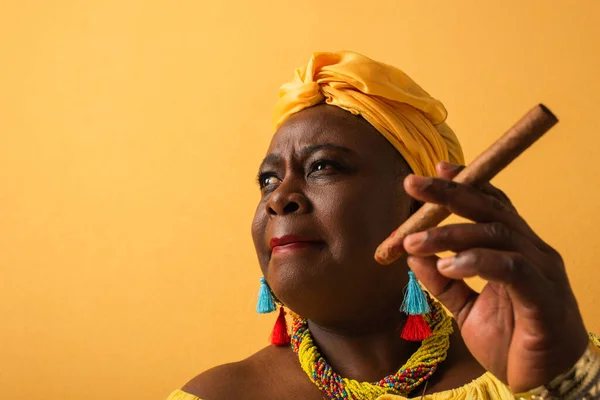 Portrait of middle aged african american woman in yellow turban holding cigar on yellow — Stock Photo