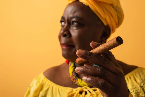 Middle aged african american woman in yellow turban and blouse holding cigar on yellow — Stock Photo