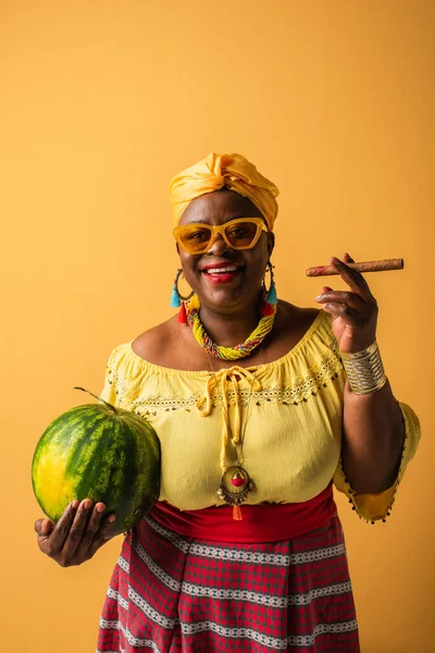 Smiling middle aged african american woman in sunglasses holding watermelon and cigar on yellow — Stock Photo