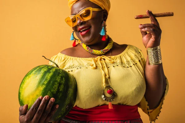 Middle aged african american woman in bright clothes and sunglasses holding watermelon and cigar on yellow — Stock Photo