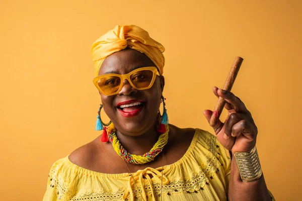Sorridente donna afroamericana di mezza età in turbante giallo e occhiali da sole che tiene il sigaro sul giallo — Foto stock