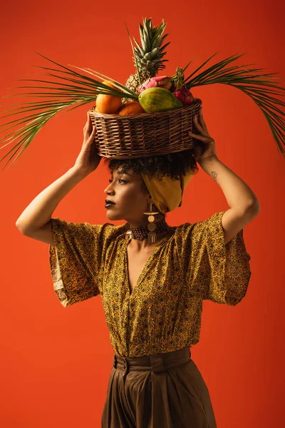 Sérieux jeune afro-américaine femme tenant panier avec des fruits exotiques sur la tête sur rouge — Photo de stock