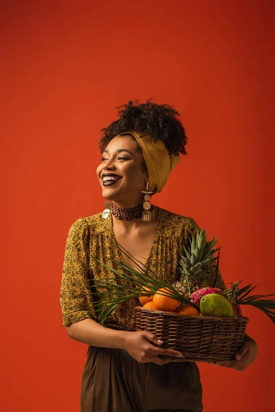 Smiling young african american woman holding basket with exotic fruits on red — Stock Photo