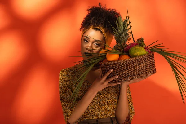 Young adult african american woman holding basket with exotic fruits near head on orange — Stock Photo