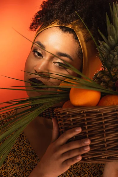 Young adult african american woman holding basket with exotic fruits near face on orange — Stock Photo
