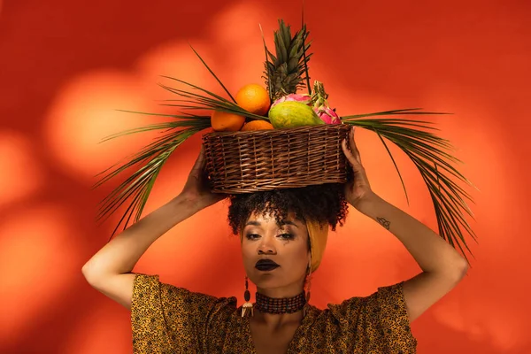 Mujer afroamericana adulta joven sosteniendo cesta con frutas exóticas en la cabeza en naranja - foto de stock