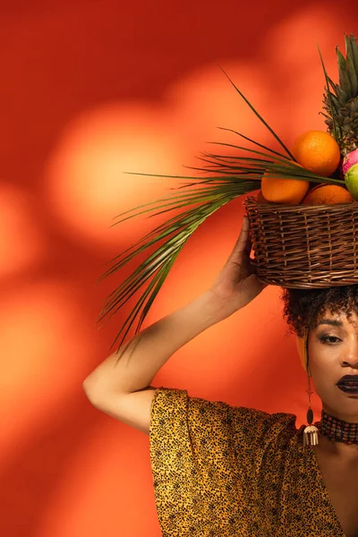 Partial view of young african american woman holding basket with exotic fruits on head on orange — Stock Photo