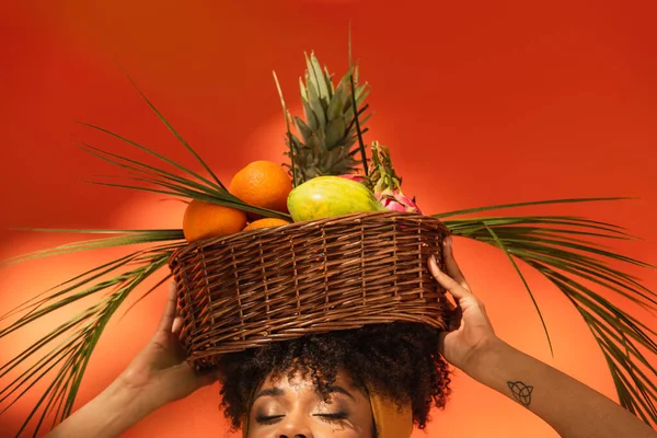 Vue partielle de jeune femme afro-américaine avec les yeux fermés tenant panier avec des fruits exotiques sur la tête sur orange — Photo de stock