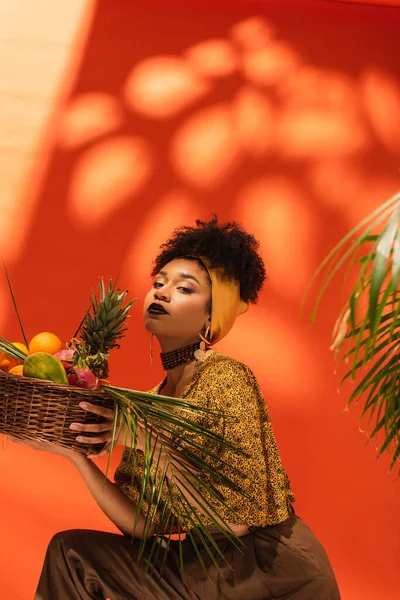 Jeune femme afro-américaine tenant panier avec des fruits exotiques et regardant la caméra sur orange — Photo de stock