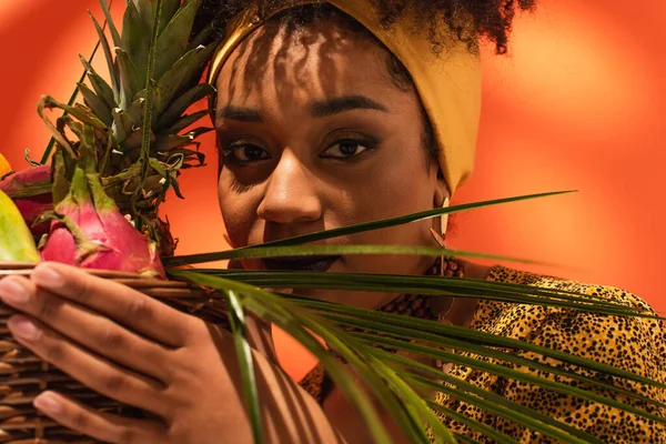 Крупный план молодой африканской женщины, держащей корзину с экзотическими фруктами рядом с лицом на апельсине — стоковое фото