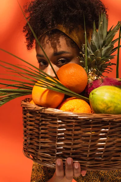 Young african american woman covering face with exotic fruits in basket on orange — Stock Photo