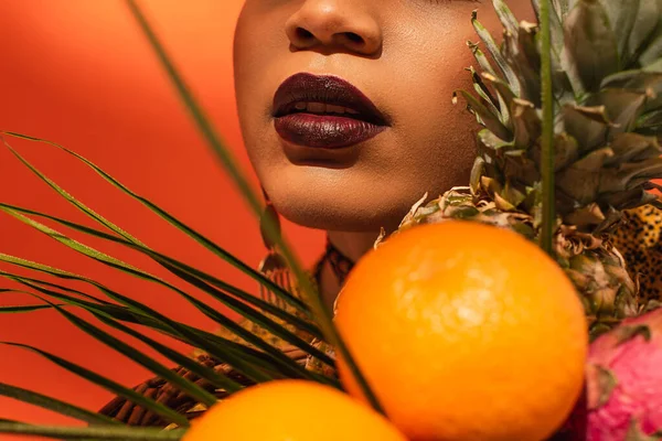 Vista de cerca de frutas y labios con lápiz labial en la cara de la joven mujer afroamericana en naranja - foto de stock
