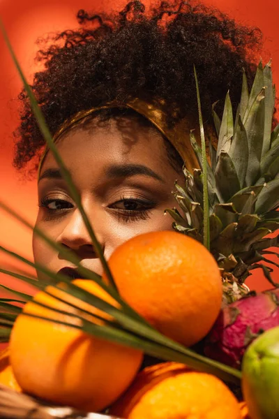 Joven afroamericana mujer ocultando la cara detrás de frutas con hojas de palma en naranja - foto de stock