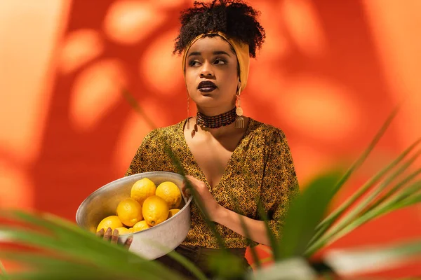 Donna afroamericana in possesso di ciotola di metallo con limoni su arancione — Foto stock