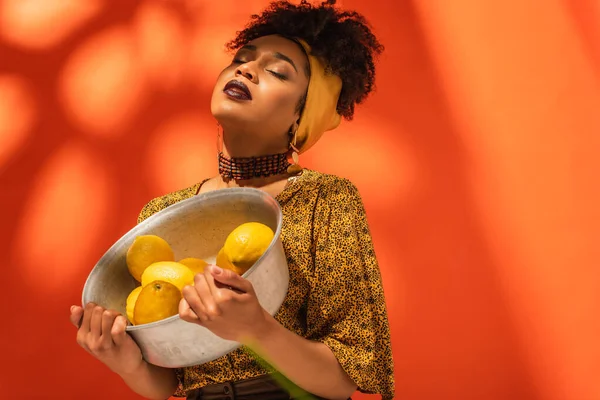 Afroamerikanerin in Bluse und Kopftuch hält Metallschale mit Zitronen auf Orange — Stockfoto