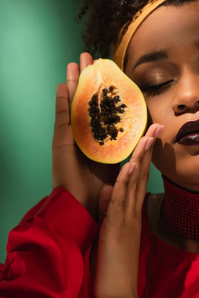 Partial view of young african american woman with closed eyes holding half cut of ripe papaya near face on green — Stock Photo