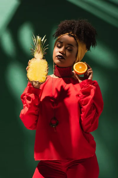 Young african american woman with closed eyes holding half cut of pineapple and orange on green — Stock Photo