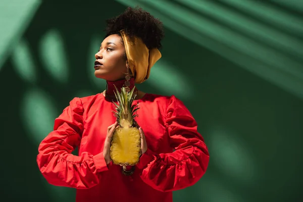Serious young african american woman holding half cut of pineapple in hands on green — Stock Photo