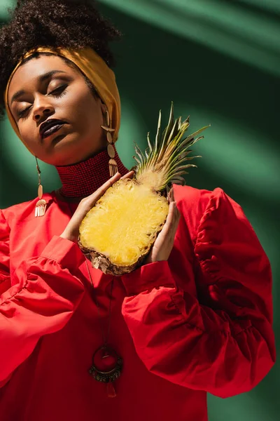 Young african american woman in red blouse holding half cut of pineapple in hands with closed eyes on green — Stock Photo