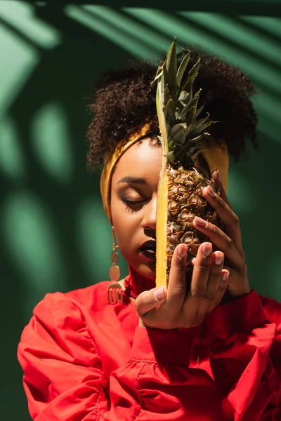 Junge afrikanisch-amerikanische Frau mit halbem Schnitt Ananas nahe Gesicht auf grün — Stockfoto