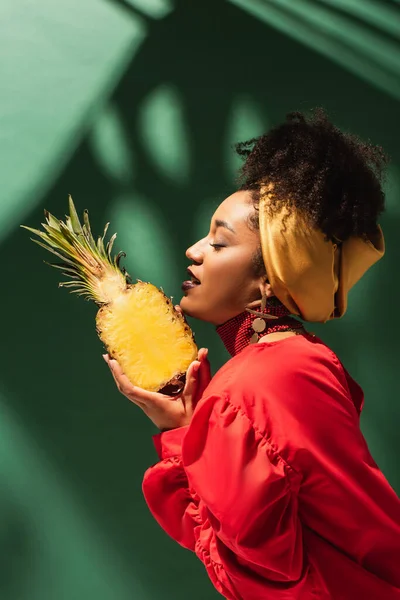 Vue latérale de la jeune femme afro-américaine avec les yeux fermés tenant demi-coupe d'ananas dans les mains sur vert — Photo de stock