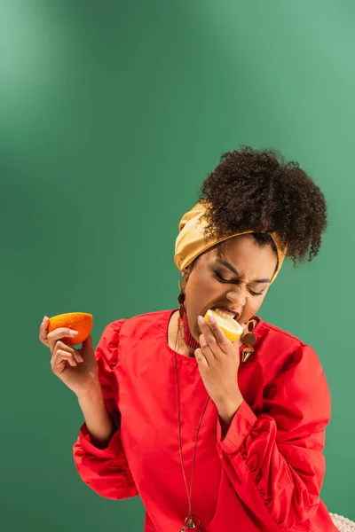 Young african american woman holding orange and eating half cut of lemon on green — Stock Photo