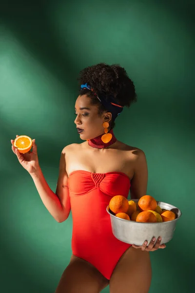 African american woman in red swimsuit holding cut orange and metal bowl with fruits on green — Stock Photo