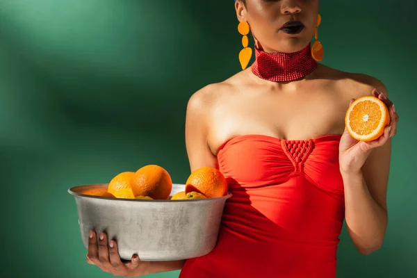 Vista parcial de mujer afroamericana en traje de baño rojo sosteniendo corte naranja y cuenco de metal con frutas en verde - foto de stock