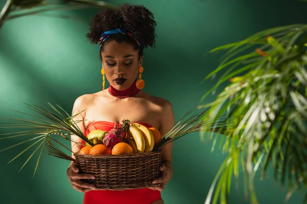 Joven afroamericana mujer mirando cesta con frutas exóticas en verde - foto de stock