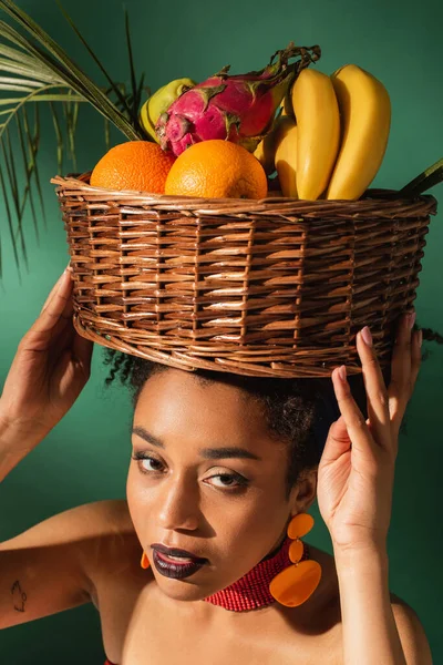 High angle view of pretty young african american woman holding basket with exotic fruits on head on green — Stock Photo