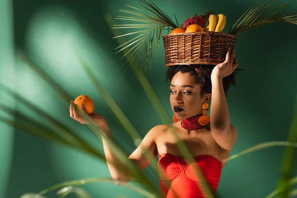 Jovem afro-americana segurando laranja e cesta com frutas exóticas na cabeça atrás de folhas de palma desfocadas no verde — Fotografia de Stock