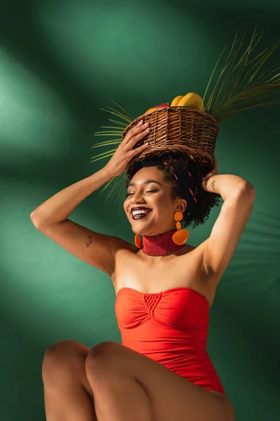 Усміхнена молода афроамериканка в червоному купальнику сидить з кошиком фруктів на голові на зеленому — стокове фото
