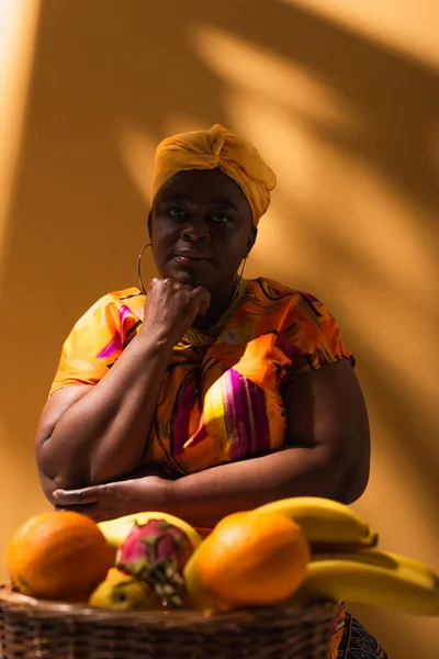 Thoughtful middle aged african american saleswoman sitting near fruits with hand near face on orange — Stock Photo