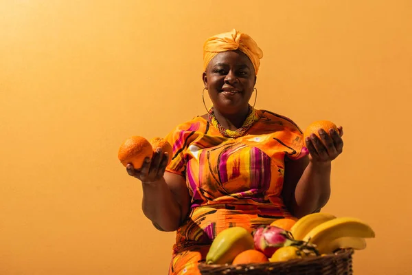 Smiling middle aged african american saleswoman sitting near fruits on orange — Stock Photo