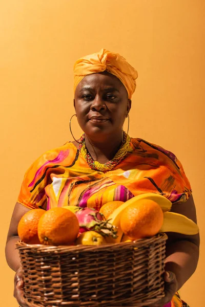 Afrikanische Amerikanerin mittleren Alters in hellem Kleid mit Korb mit exotischen Früchten auf Orange — Stockfoto