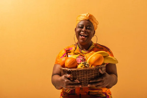 Smiling middle aged african american woman holding basket with fruits on orange — Stock Photo