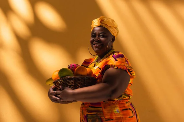 Cheerful middle aged african american woman holding basket with exotic fruits and looking at camera on orange — Stock Photo