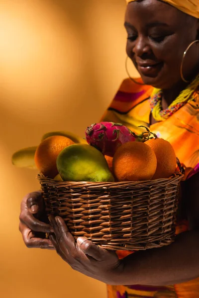 Donna afroamericana di mezza età in abito luminoso cesto con frutta esotica su arancione — Foto stock
