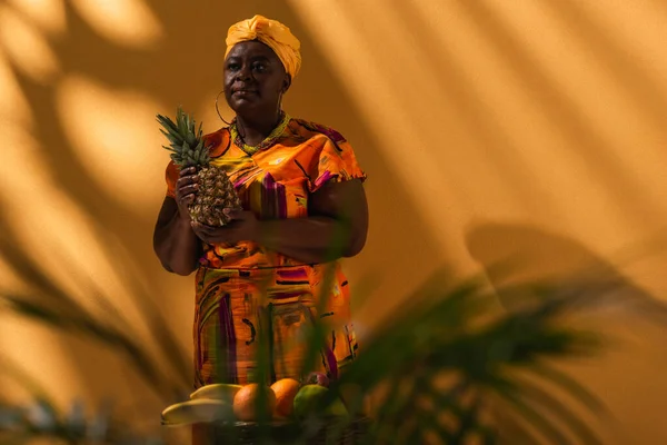Vendedora afro-americana de meia idade de pé com abacaxi perto de frutas em laranja — Fotografia de Stock