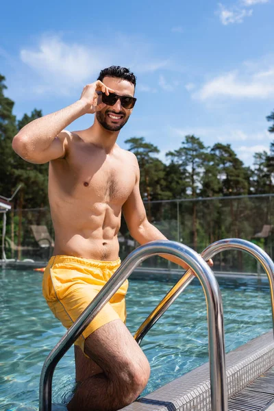 Усміхнений арабський чоловік в сонцезахисних окулярах стоїть біля басейну — стокове фото