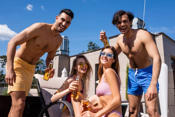 Amigos multiétnicos em roupa de banho segurando garrafas de cerveja ao ar livre — Fotografia de Stock