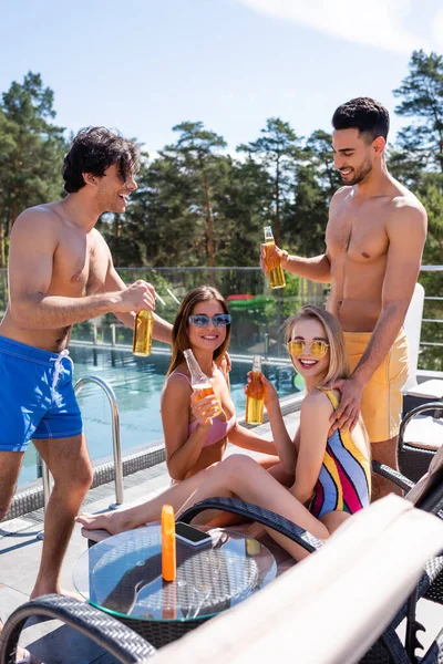 Mulheres sorridentes em roupa de banho segurando garrafas de cerveja perto de amigos inter-raciais e piscina — Fotografia de Stock