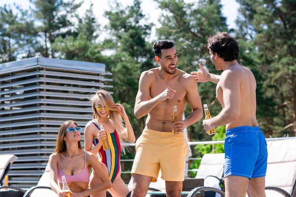 Allegro uomini multietnici con birra parlando vicino amici in costumi da bagno all'aperto — Foto stock