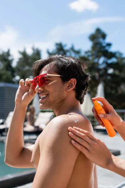 Mujer que aplica protector solar en la espalda de un amigo sonriente con gafas de sol - foto de stock