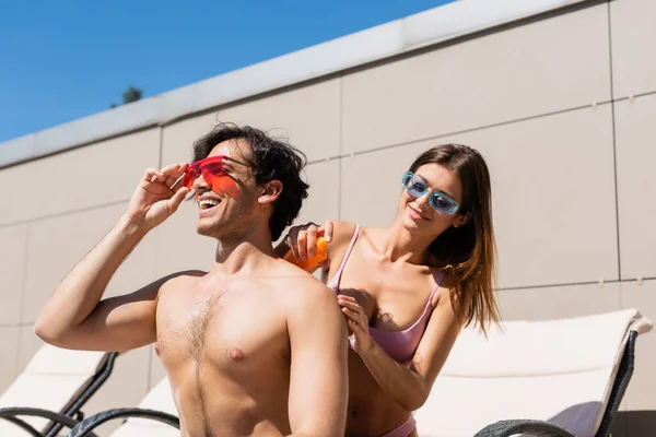 Femme souriante en maillot de bain appliquant de la crème solaire sur un ami en lunettes de soleil — Photo de stock