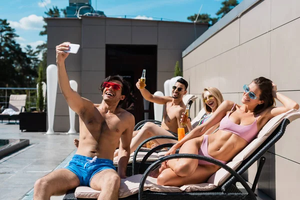 Felices amigos multiétnicos con cerveza tomando selfie en el teléfono inteligente en las tumbonas - foto de stock